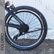 Close-up of a folded bike’s rear wheel fitted with a Schwalbe tire and the FOLDRIO Brompton Kickstand – Carbon Fibre, P and T Line, set against white tiles and concrete. Left side displays a vertical transparent FOLDRIO strip.