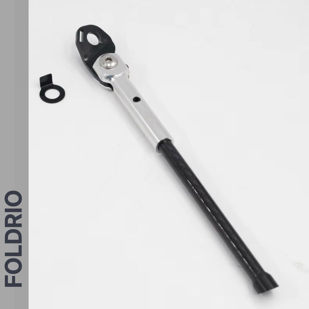The FOLDRIO Brompton Kickstand – Carbon Fibre, P and T Line is shown in silver and black with a small black accessory, displayed against a white background. The FOLDRIO logo appears vertically on a gray strip on the left edge.