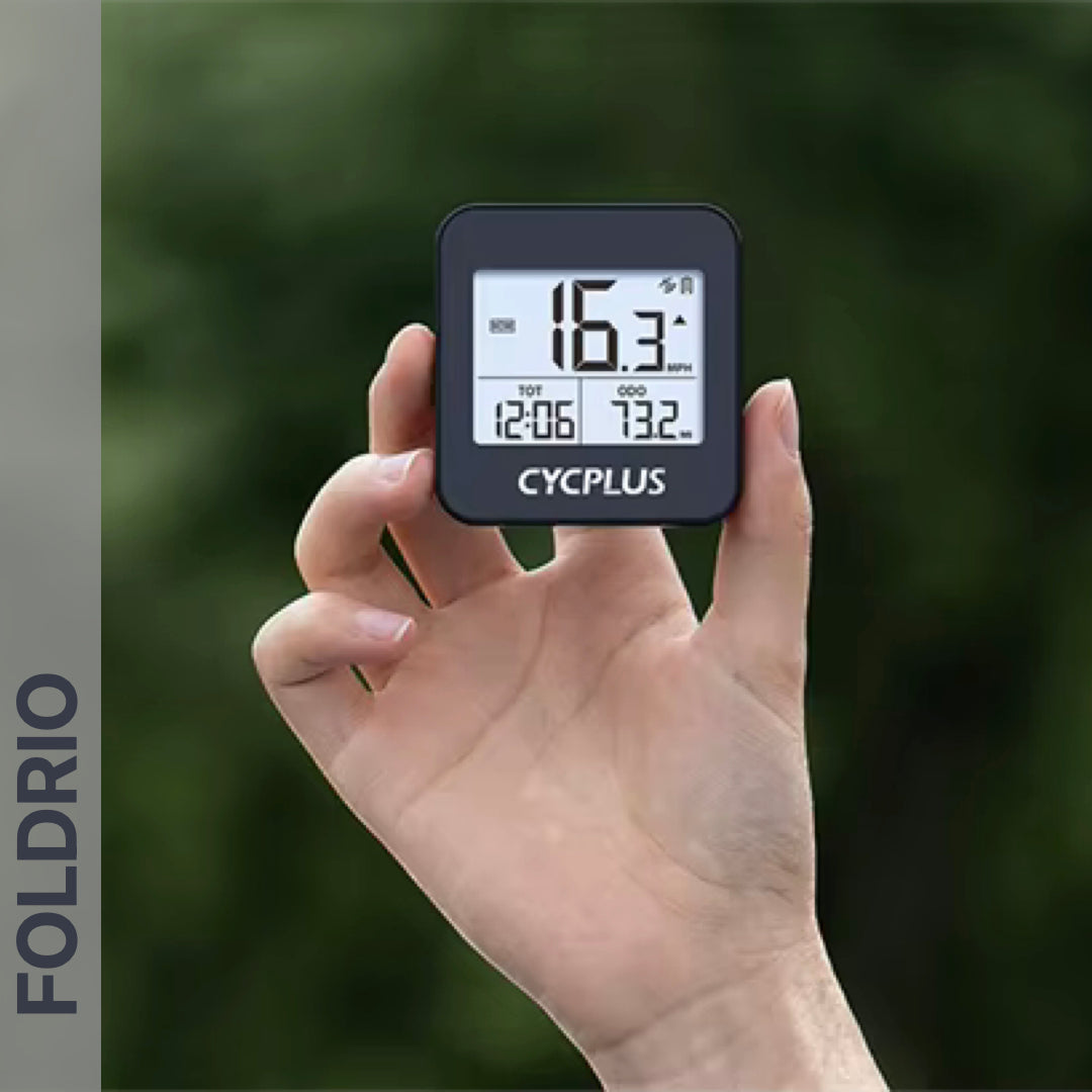 A hand holds the FOLDRIO Brompton GPS Bike Computer (wireless, IPX6, 9 data fields) with numbers and symbols on its black screen; "FOLDRIO" appears vertically left, and blurred greenery forms the background.