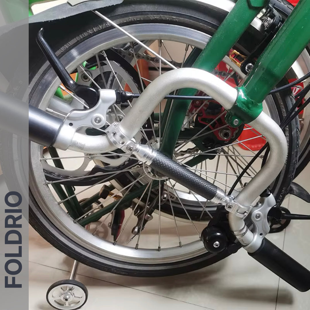 A close-up shows the FOLDRIO Brompton M Handlebar Cross Brace – Adjustable, available in 6 colours, compactly arranged on a folded green bike. The word FOLDRIO appears vertically on the left side of the image.
