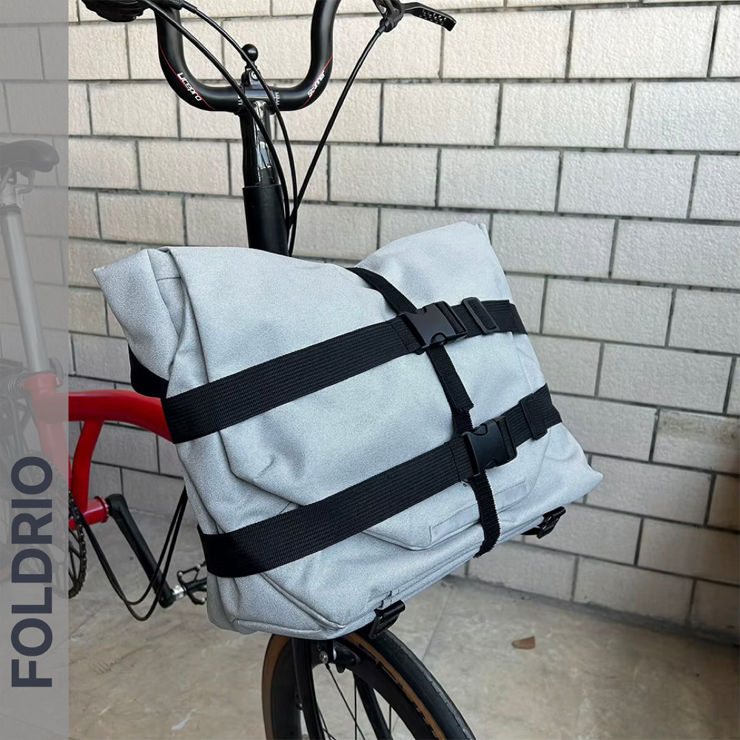 A FOLDRIO Brompton Front Bag Rack Holder with elastic straps is attached to the handlebars of a red Brompton Folding Bike, parked on a concrete floor against a white brick wall. "FOLDRIO" appears on the left side.