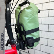 A Brompton Front Bag Rack Holder with elastic straps by FOLDRIO is securely attached to the frame of a red Brompton folding bike, which rests against a white brick wall with the FOLDRIO brand partially visible on the left.
