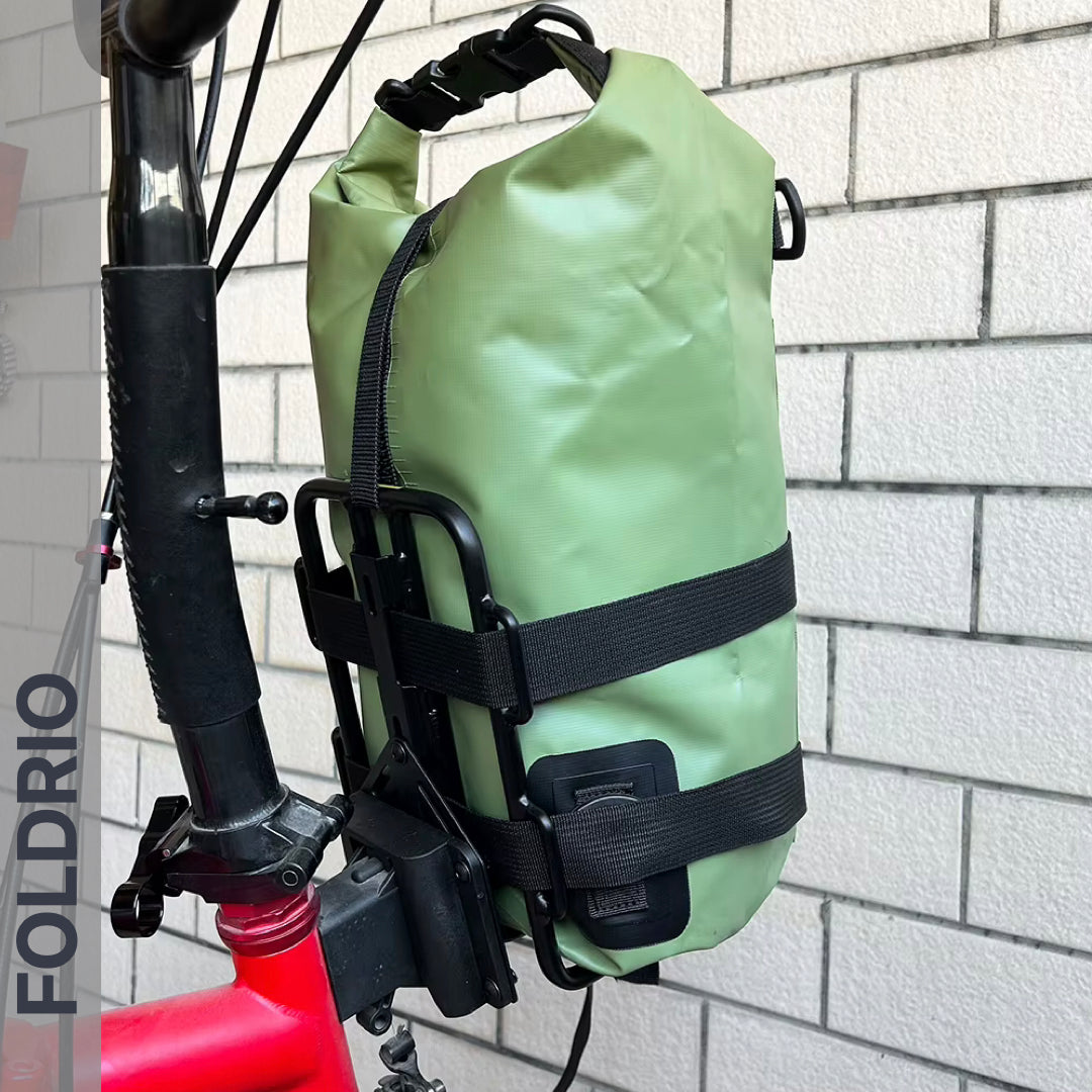 A Brompton Front Bag Rack Holder with elastic straps by FOLDRIO is securely attached to the frame of a red Brompton folding bike, which rests against a white brick wall with the FOLDRIO brand partially visible on the left.