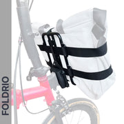 A white FOLDRIO Brompton Front Bag Rack Holder with elastic straps is mounted on a red bike frame using black straps and a black metal rack. The background is faded, and "FOLDRIO" appears vertically on the left.