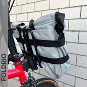 A FOLDRIO Brompton Front Bag Rack Holder with Elastic Straps is mounted on a red bike, positioned against a white brick wall. The word FOLDRIO appears vertically along the image’s left edge.