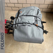 A gray backpack is secured on a bicycle's rear rack with FOLDRIO Brompton Rear Rack Elastic Straps (fits AC Line & PT Line), on a tiled floor with a brick wall behind. The word FOLDRIO appears along the left side.