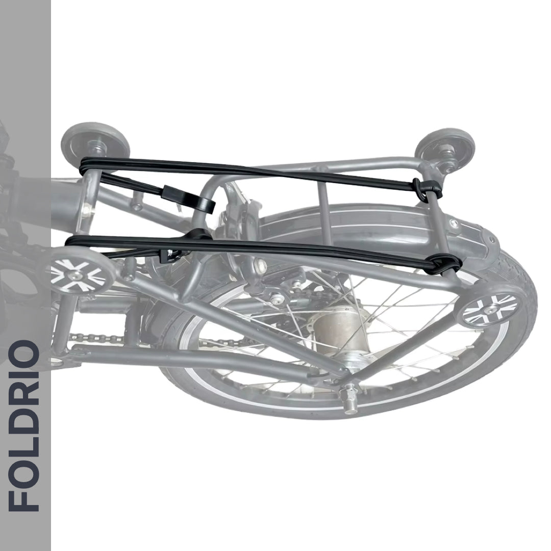 A folded bicycle is secured by Brompton Rear Rack Elastic Straps from FOLDRIO (fits AC Line & PT Line), with the word FOLDRIO displayed vertically on the left side. The wheels and frame are neatly aligned.