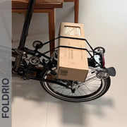 A folding bike with a beige box secured to its rear rack using FOLDRIO Brompton Rear Rack Elastic Straps for AC Line & PT Line stands indoors on white tiles near wooden chairs. "FOLDRIO" appears vertically on the left.