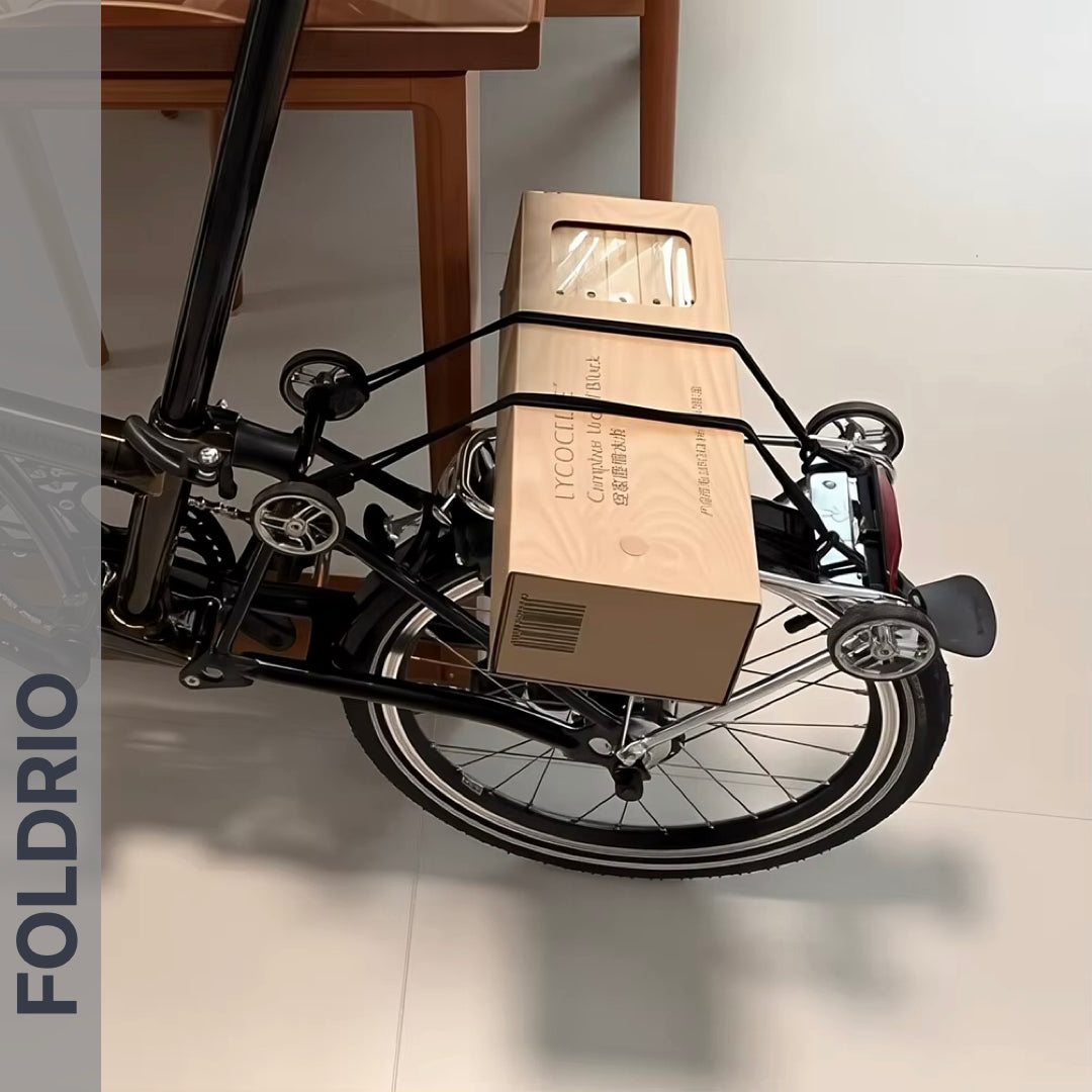 A folding bike with a beige box secured to its rear rack using FOLDRIO Brompton Rear Rack Elastic Straps for AC Line & PT Line stands indoors on white tiles near wooden chairs. "FOLDRIO" appears vertically on the left.