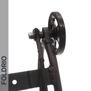 Close-up of FOLDRIO Brompton Rear Rack Elastic Straps (fits AC Line & PT Line) on black pulley wheel attached to metal frame, with bolt, cable, and bike rack strap. “FOLDRIO” appears vertically on a gray stripe; background is white.