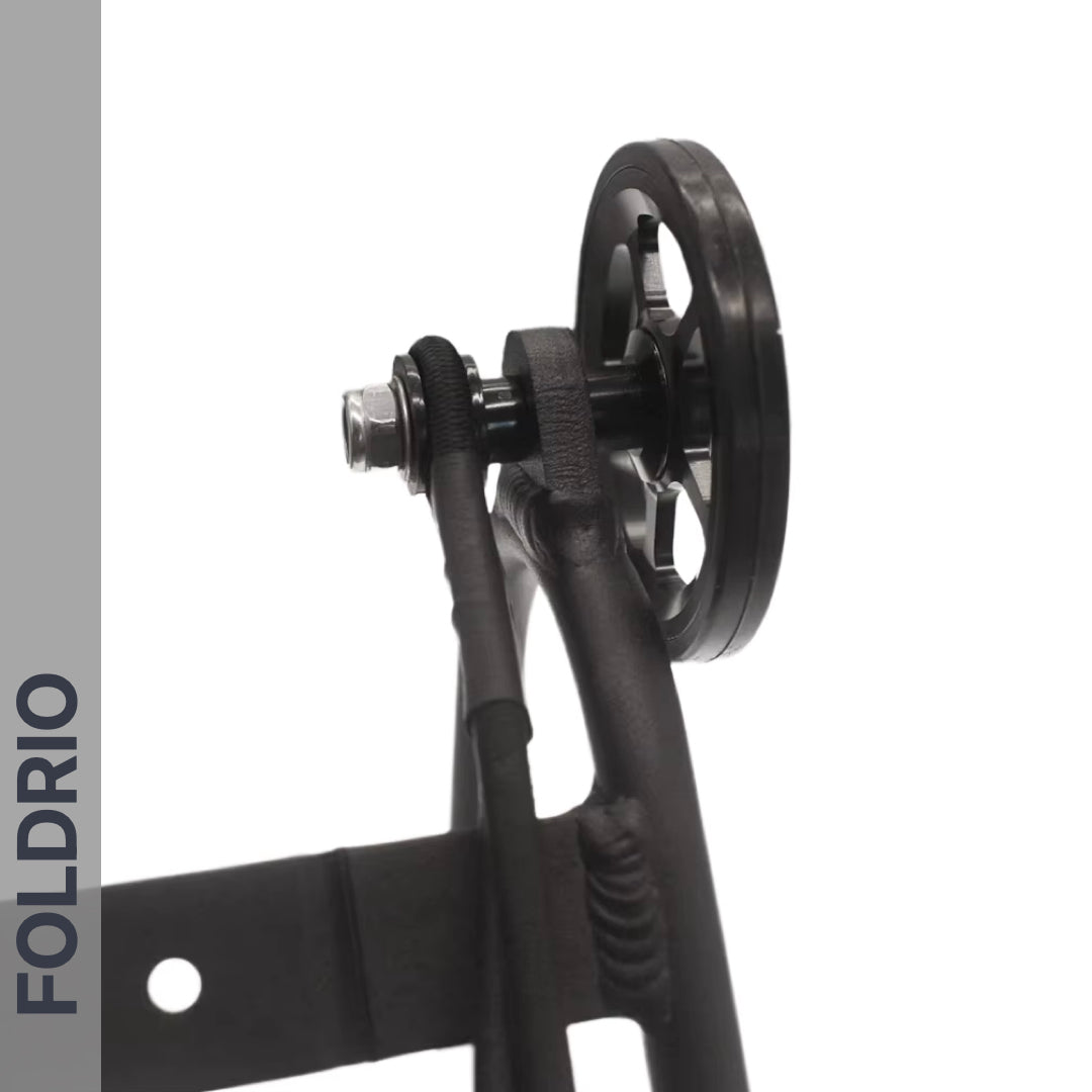 Close-up of FOLDRIO Brompton Rear Rack Elastic Straps (fits AC Line & PT Line) on black pulley wheel attached to metal frame, with bolt, cable, and bike rack strap. “FOLDRIO” appears vertically on a gray stripe; background is white.
