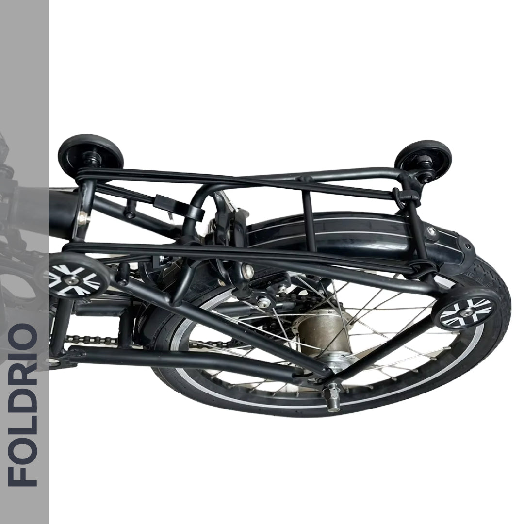 A black folding bicycle, viewed from above in its compact folded state, is secured with Brompton Rear Rack Elastic Straps by FOLDRIO. The brand name FOLDRIO is displayed vertically on the left side of the image.