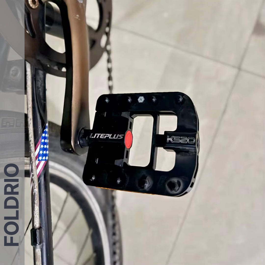 Close-up of a black Brompton Foldable Flat Pedal – Lightweight Aluminium Upgrade attached to a bike. Its design is highlighted against a tiled floor, with FOLDRIO displayed vertically on the left side of the image.