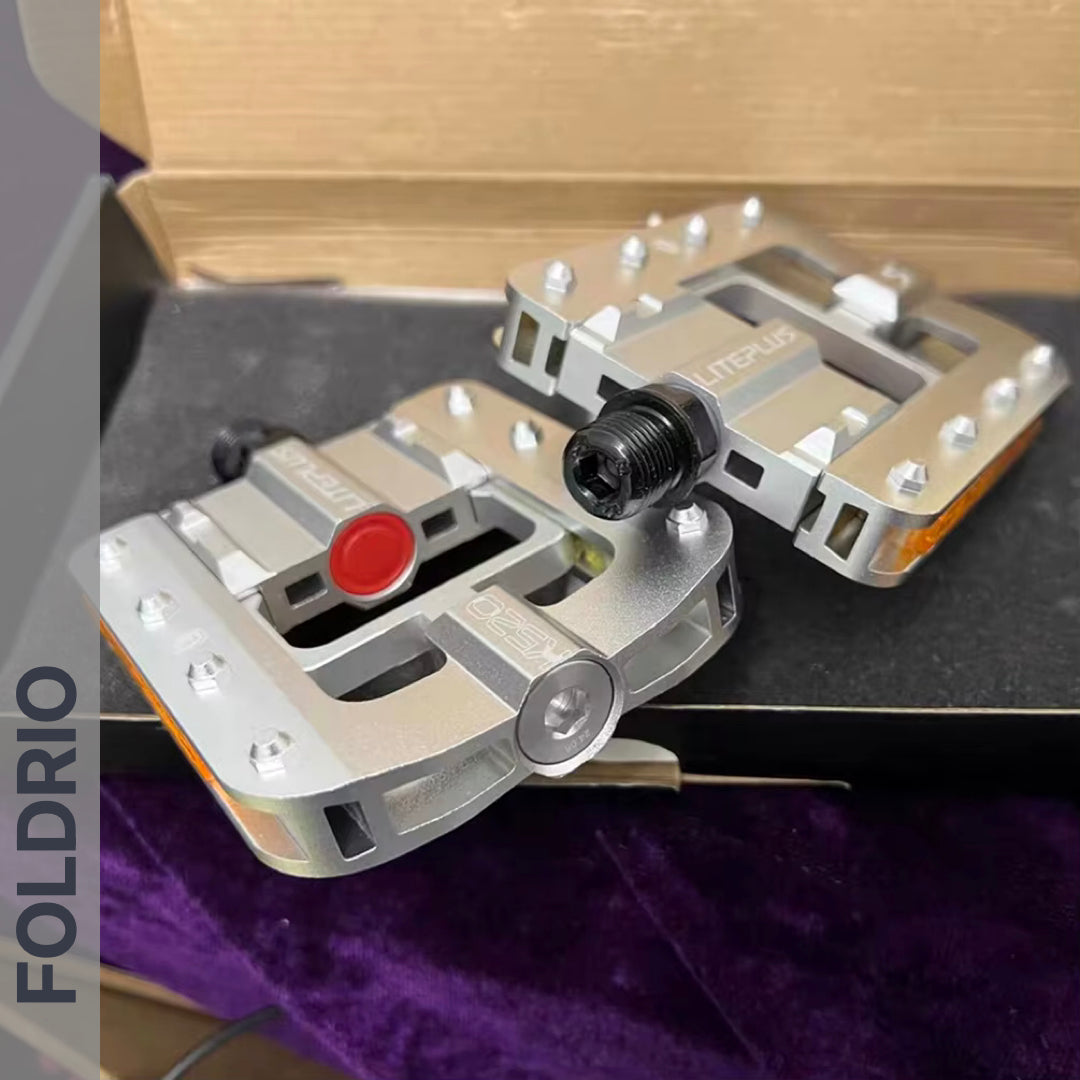 Two Brompton Foldable Flat Pedals – Lightweight Aluminium Upgrade, each with reflective strips and visible branding, rest on a black and purple surface. A cardboard box is behind them; one pedal features a red circle, the other a silver circle at the end.