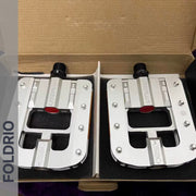 A pair of Brompton Foldable Flat Pedals – Lightweight Aluminium Upgrade in silver with black axles, ideal for folding bikes, packaged in an open box with black foam padding. The word FOLDRIO is on the left edge.