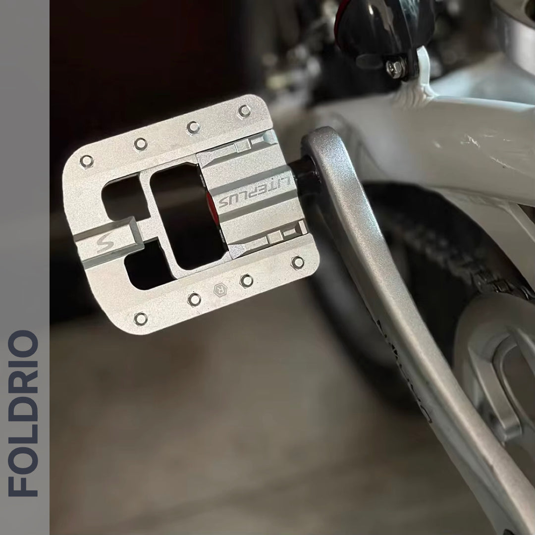 Close-up of a Brompton Foldable Flat Pedal—lightweight aluminium—mounted on a silver crank arm. A white bike frame and rear tire are in the background. The word FOLDRIO is shown vertically on the left margin.