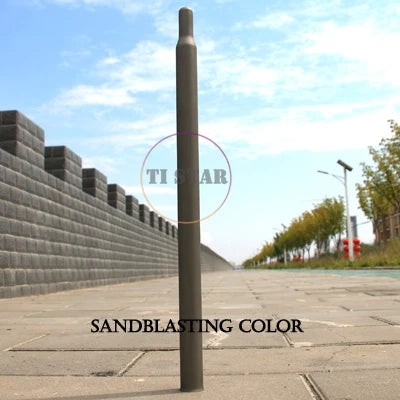 A Brompton Titanium Seatpost – Multicolour Edition by FOLDRIO stands upright on a paved walkway, bordered by a stone wall and trees under a blue sky. The text SANDBLASTING COLOR appears at the bottom.